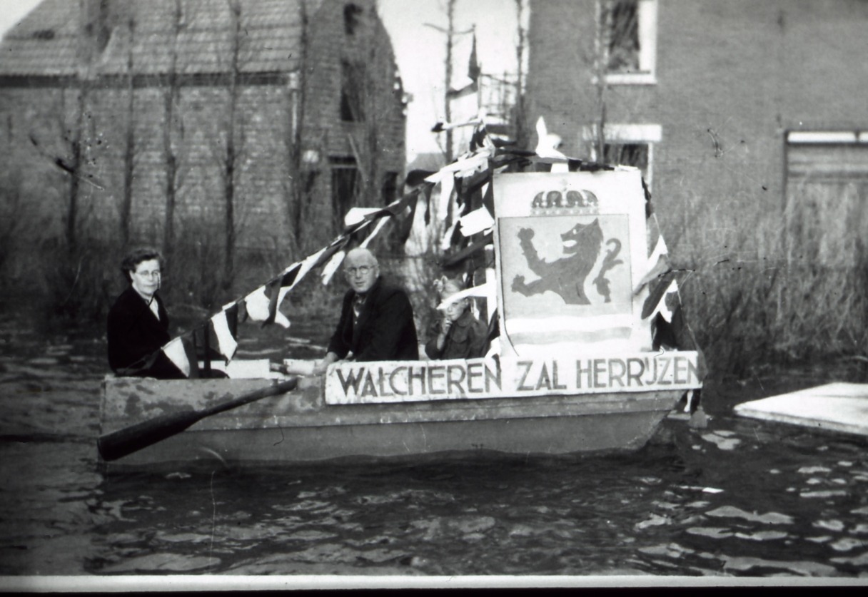 Zwart-witte foto van drie personen in een bootje door een straat varend tijdens de inundatie van Walcheren. Op dit bootje zijn vlaggen geplaatst en de tekst "Walcheren zal herrijzen".