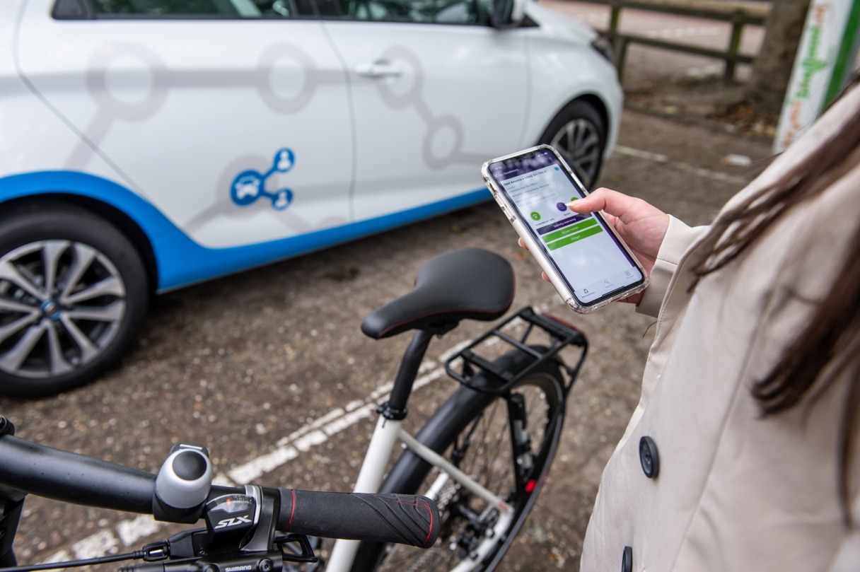 Vrouw naast fiets en deelauto gebruikt app op mobiele telefoon. Beeld: Addvision SD Op Weg