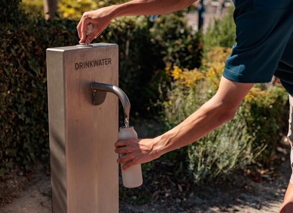 Close-up van een persoon die zijn waterfles vult bij een drinkwatertappunt in Kamperland.