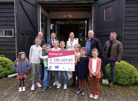 Gedeputeerde Jo-Annes de Bat samen met wethouder Joan Veldhuizen en 9 kinderen en 3 volwassen. De kinderen houden een bord vast met daarop de tekst: 'Bedrag van: 106.179,85'.