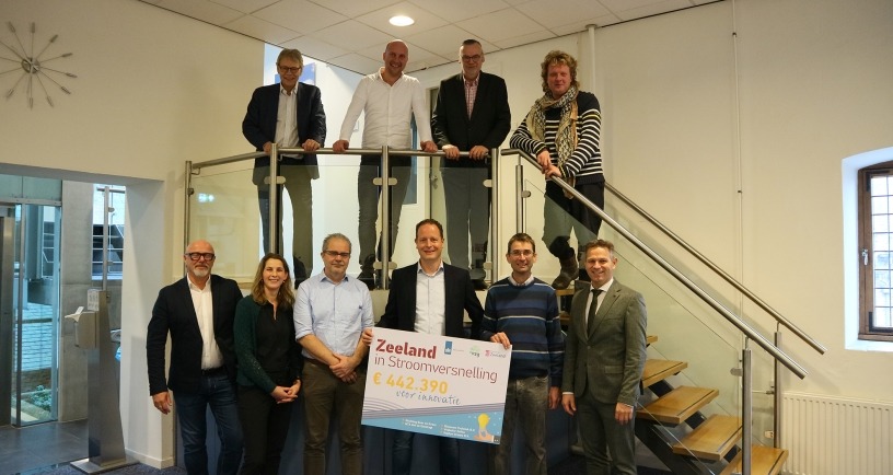 Een groepsfoto van subsidieontvangers van Zeeland in Stroomversnelling-subsidie. Vier mensen staan bovenop de trap. Een zestal staan onderaan de trap en houden een foambord vast met daarop 'Zeeland in Stroomversnelling €442.390 voor innovatie'. 