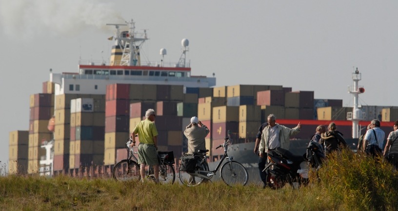Over de Westerschelde vaart een enorm containerschip. Toeschouwers met fiets op de dijk bekijken het schip.