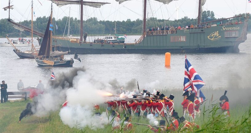Engelse soldaten schieten. Op achtergrond driemaster en diverse zeilschepen.